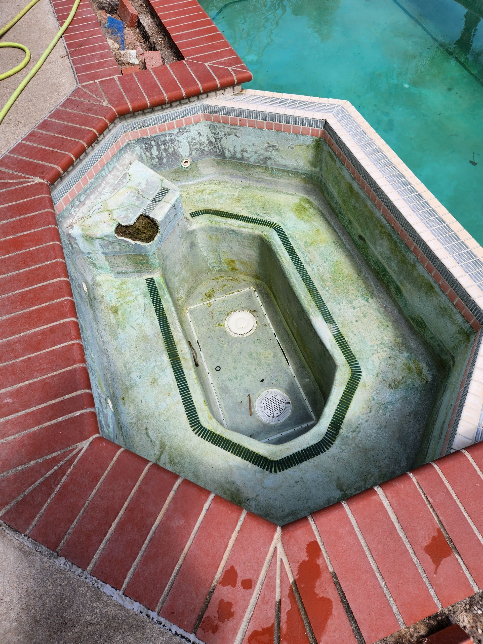A concrete spa with green algae buildup surrounded by brick. The spa is next to a pool.