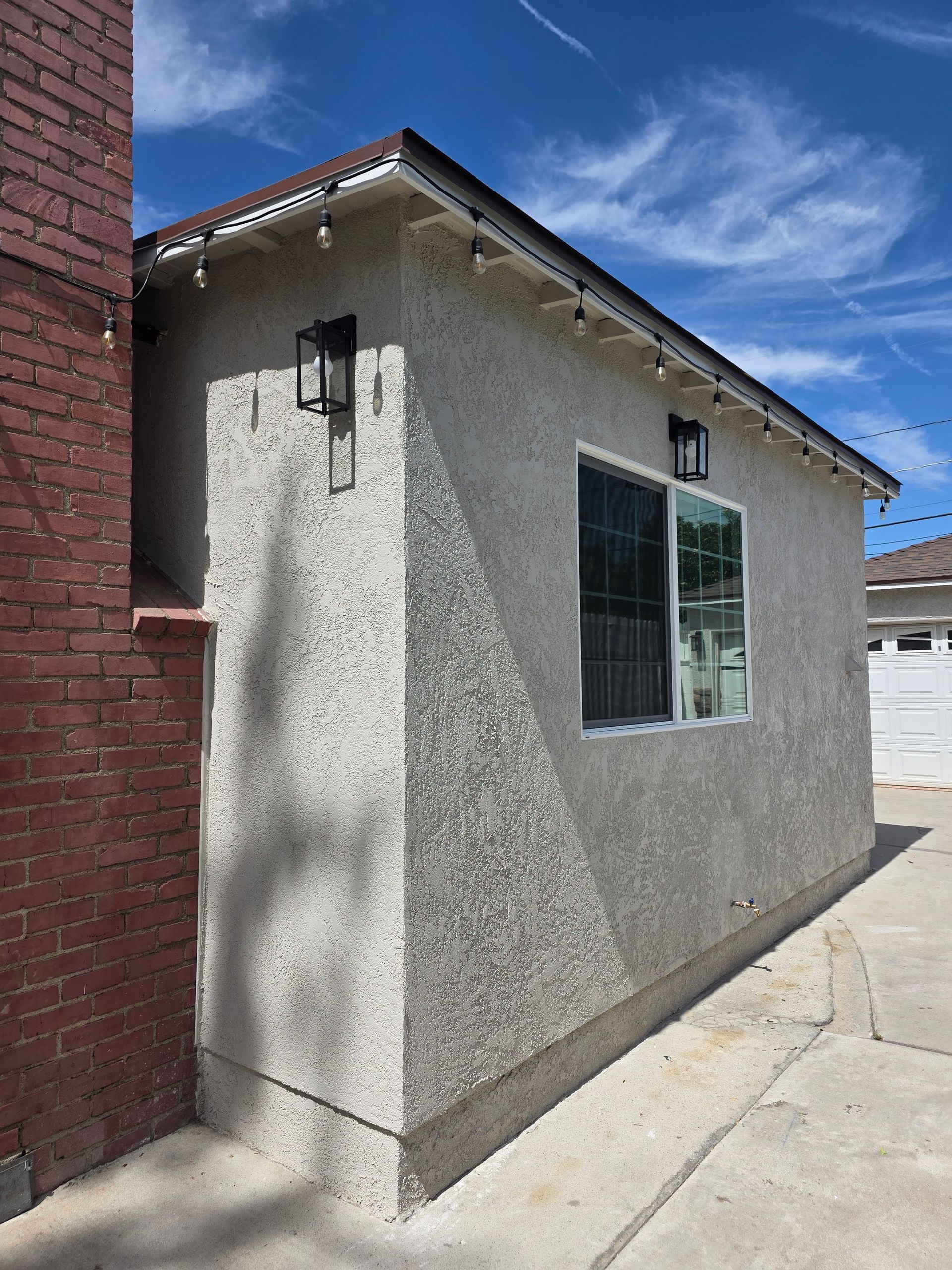 Small stucco building with a window, two wall-mounted lights, and brick chimney on a sunny day.