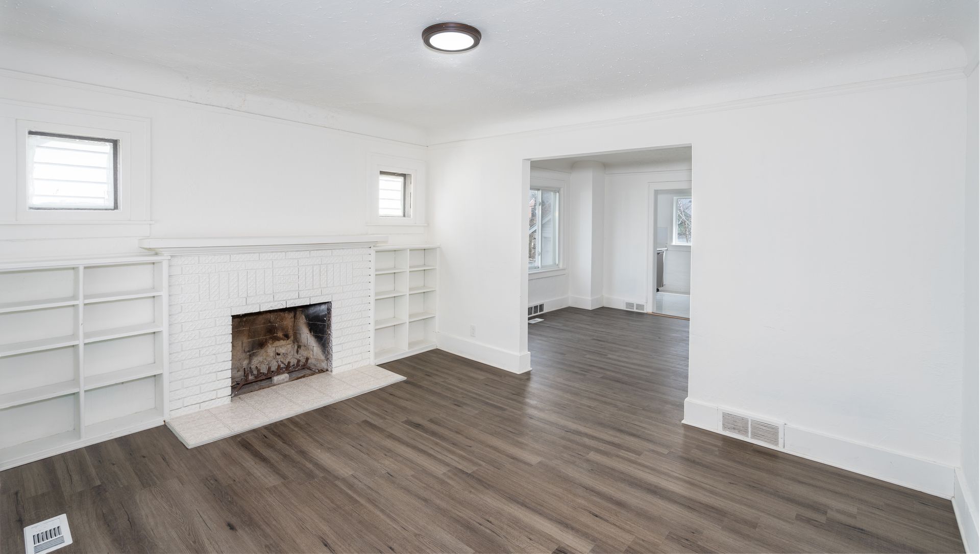 Living room with fireplace, built-in shelves, white walls, dark wood floor, and an open doorway to another room.