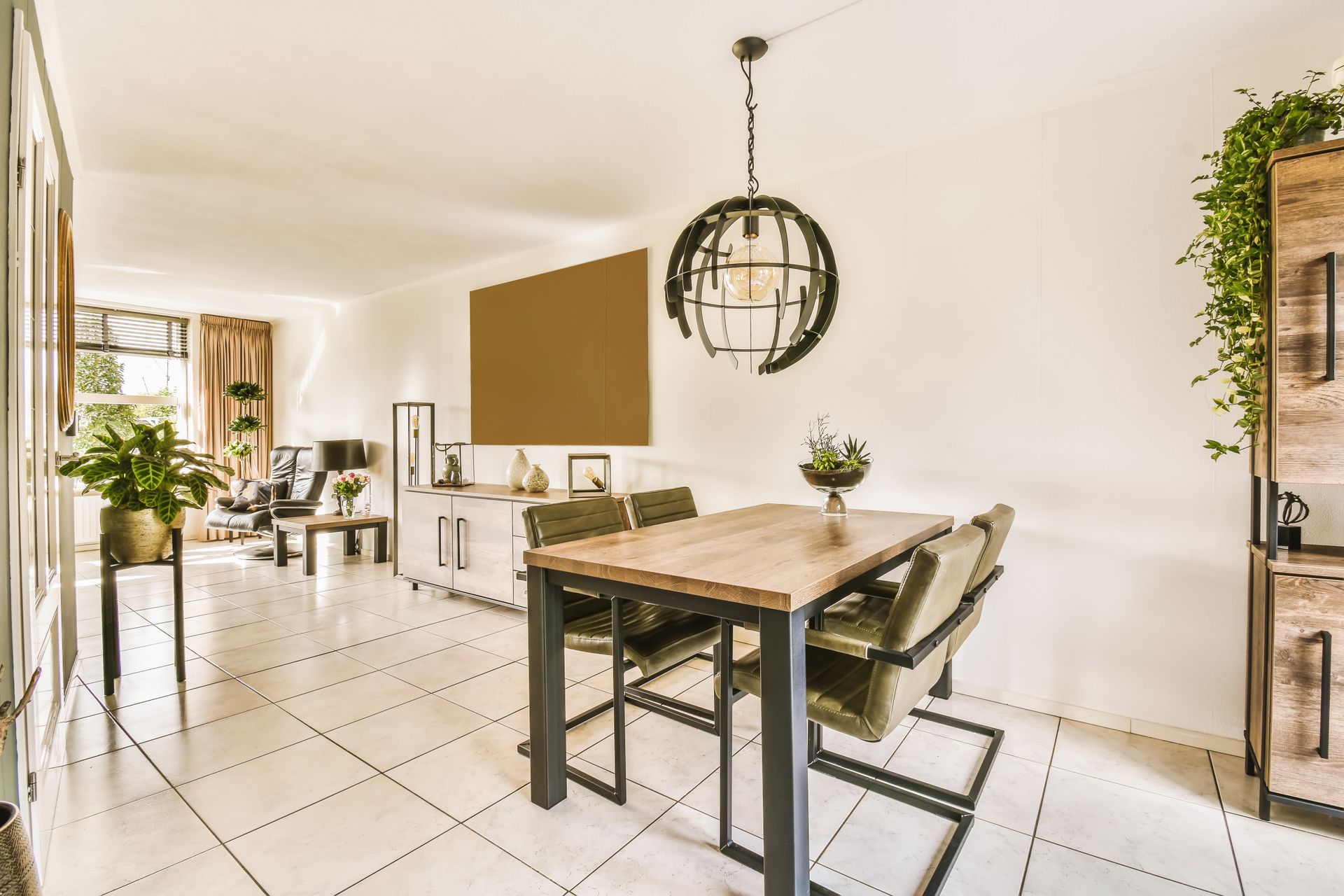 Dining area with wooden table, metal legs, and green chairs. Overhead globe light. White tile floors, bright room.