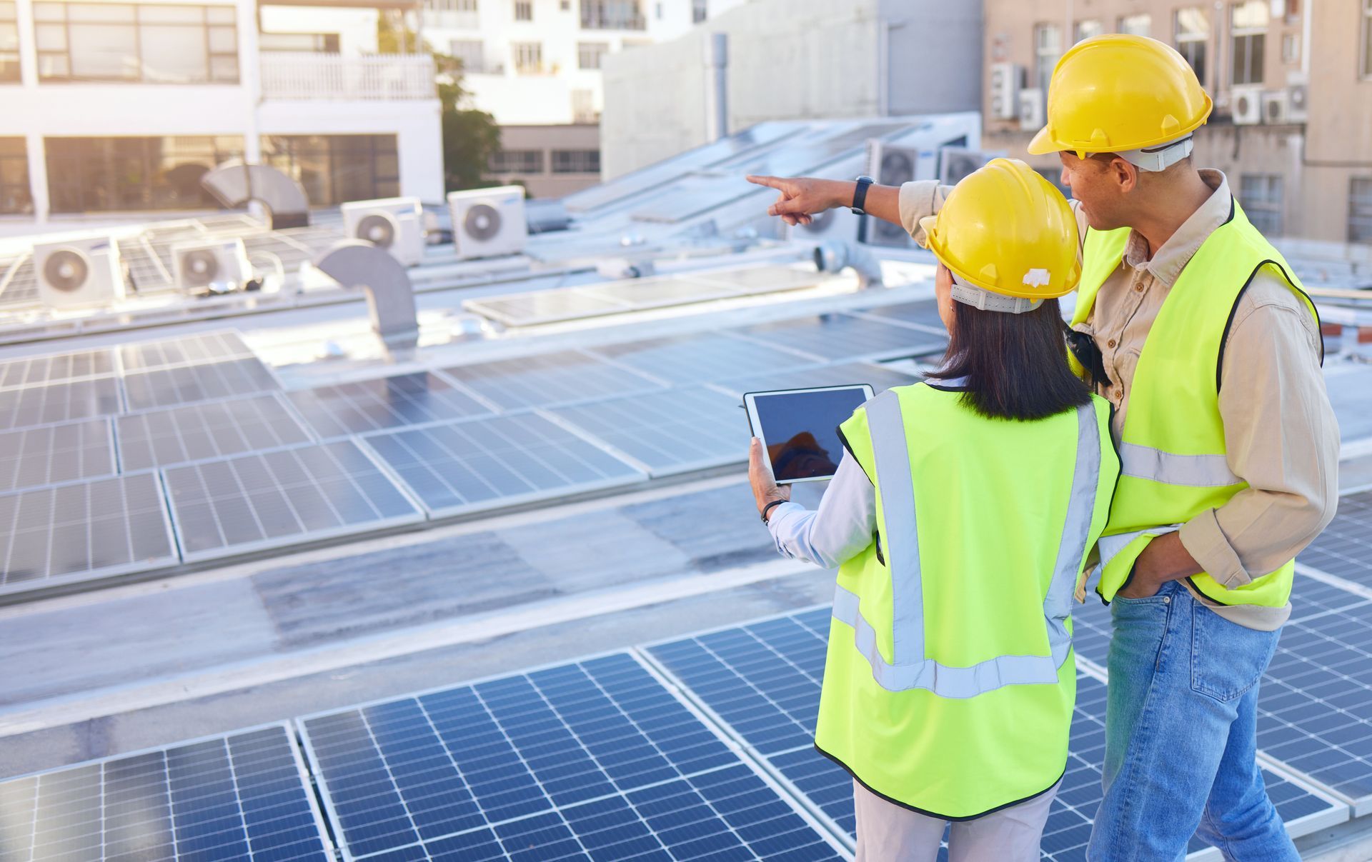 Two people in safety vests and hard hats on a rooftop with solar panels, one pointing, the other holding a tablet.