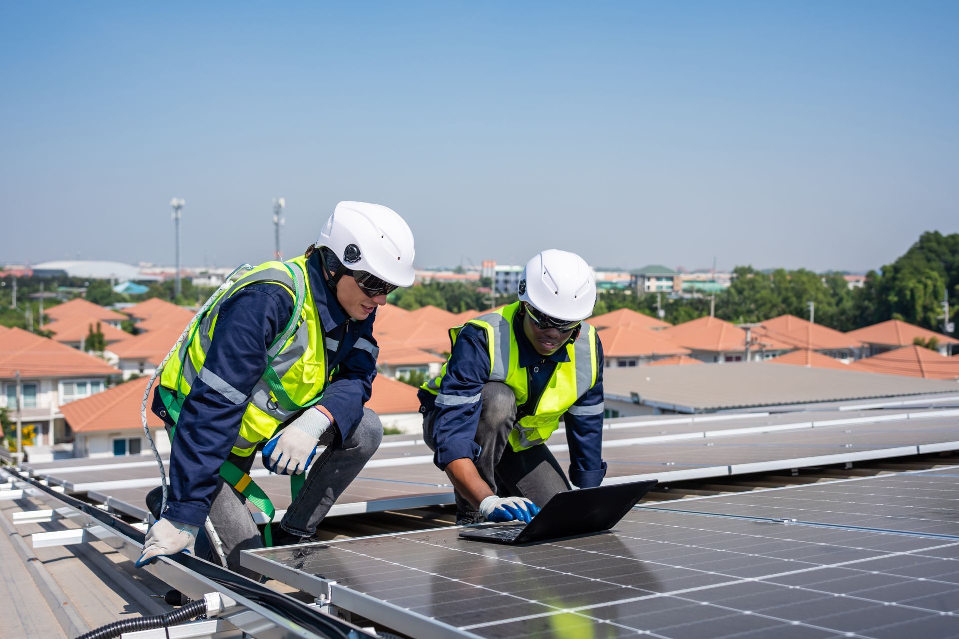 Two workers in safety gear inspect solar panels on a rooftop using a laptop.