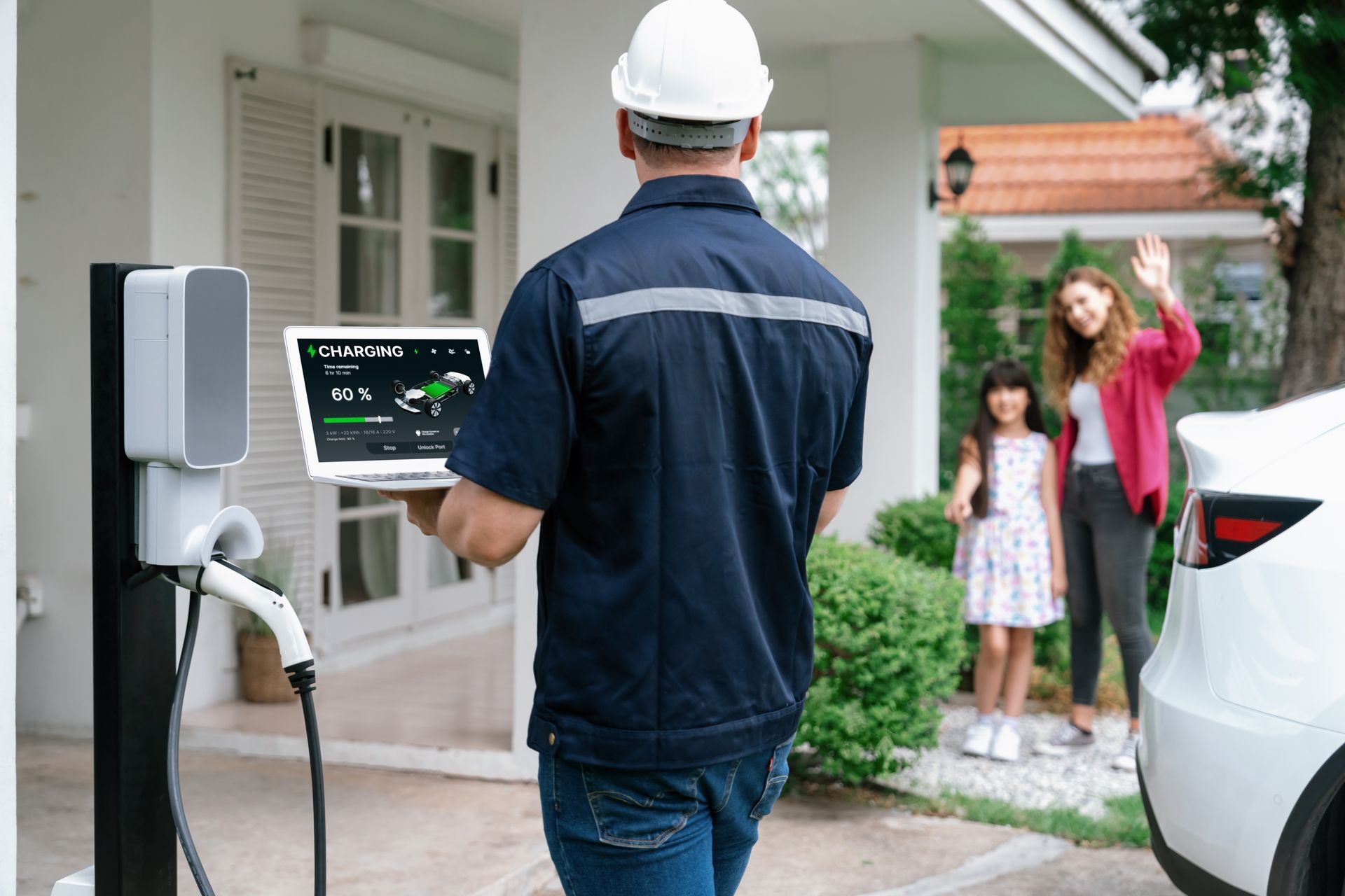 Electrician inspects EV charger with laptop as family waves from doorway.