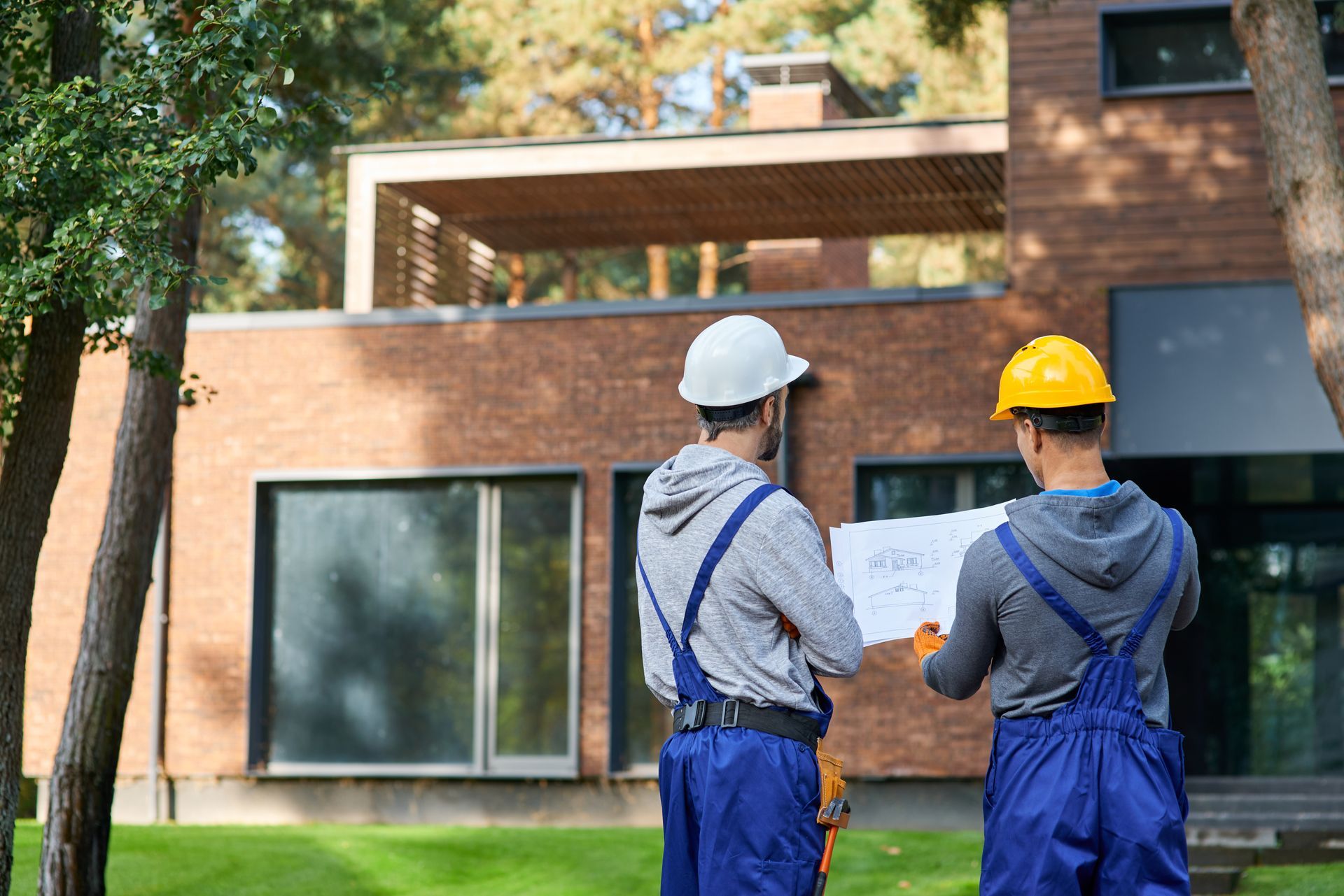 Two construction workers in blue overalls and hard hats reviewing plans outside a brick house.