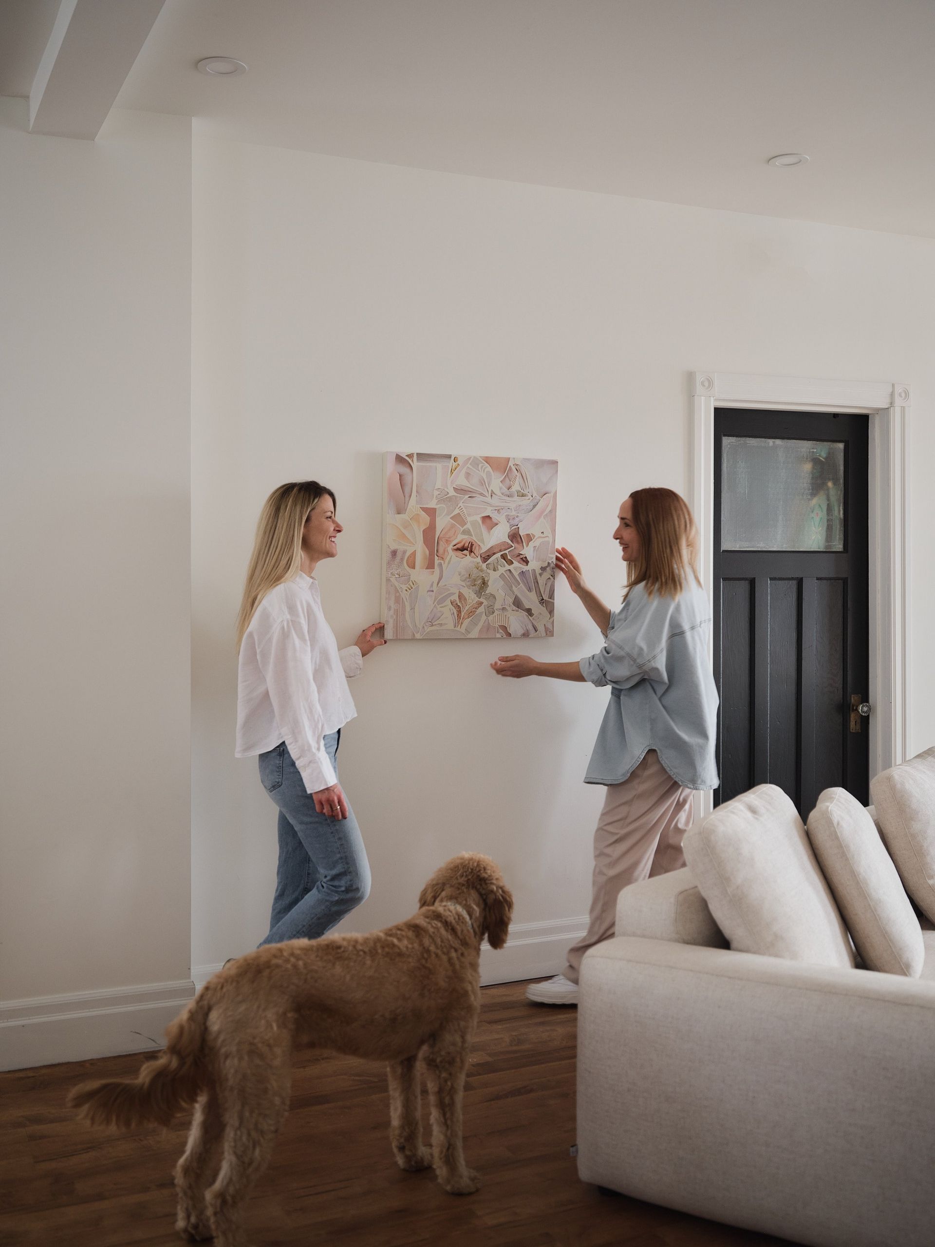 Deux femmes et un chien se tiennent dans un salon et regardent un tableau accroché au mur.