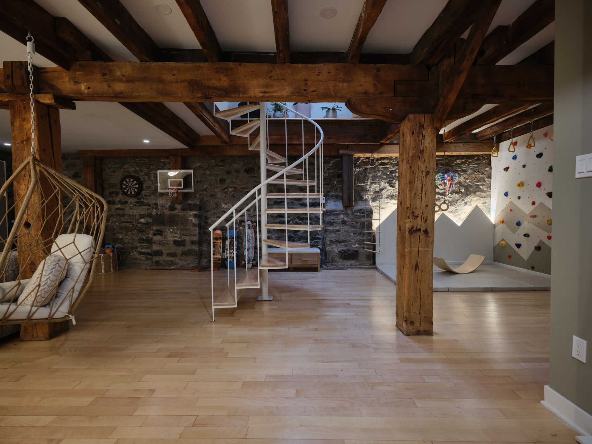 Un sous-sol avec poutres en bois, un escalier en colimaçon, des murs en pierre, un fauteuil suspendu et un mur d'escalade pour enfants.