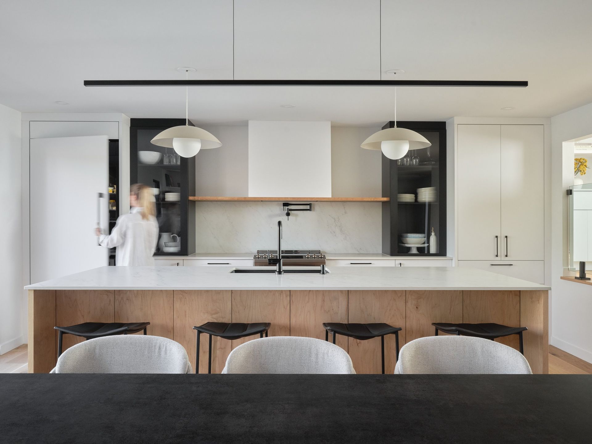 Une cuisine avec un grand îlot et des tabourets et une femme debout en arrière-plan.
