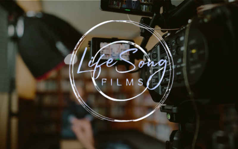 Close-up of a video camera with the Life Song Films logo overlaid. A blurred person is in the background.