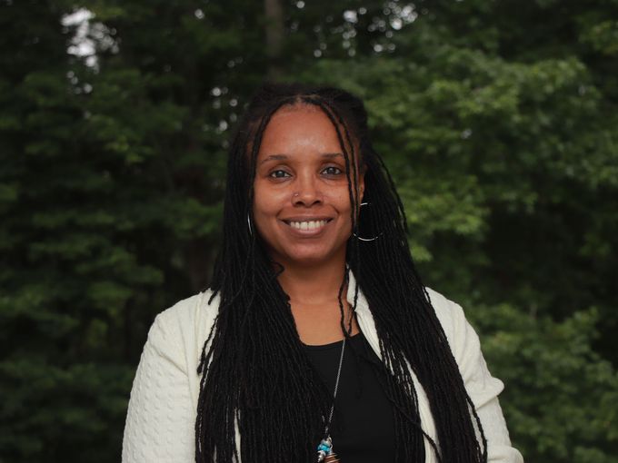 Woman with long braids smiles, wearing a white jacket in front of green trees.