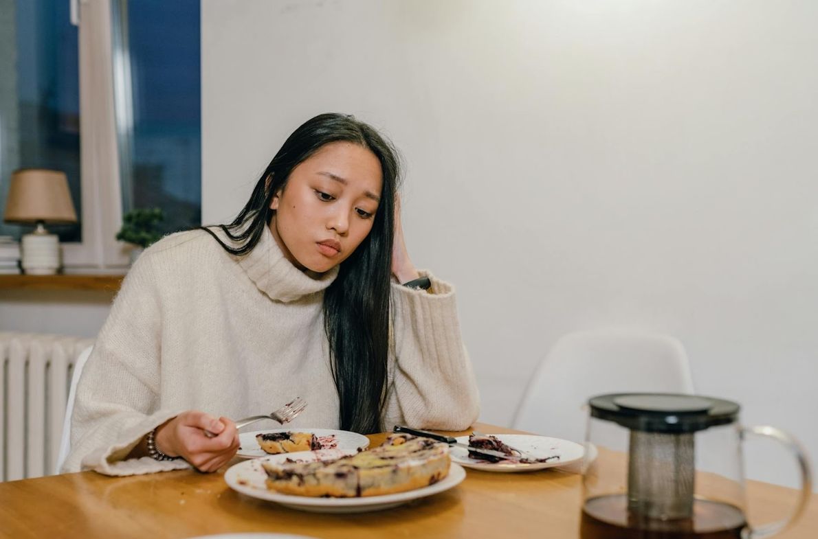 Young woman in a cream turtleneck sweater looking down at a partially eaten blueberry cake on a wood