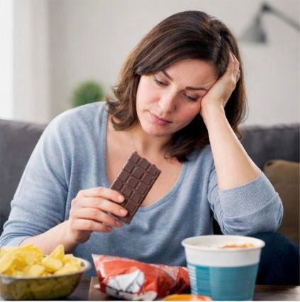 Woman looking at chocolate bar, surrounded by snacks, appearing sad.