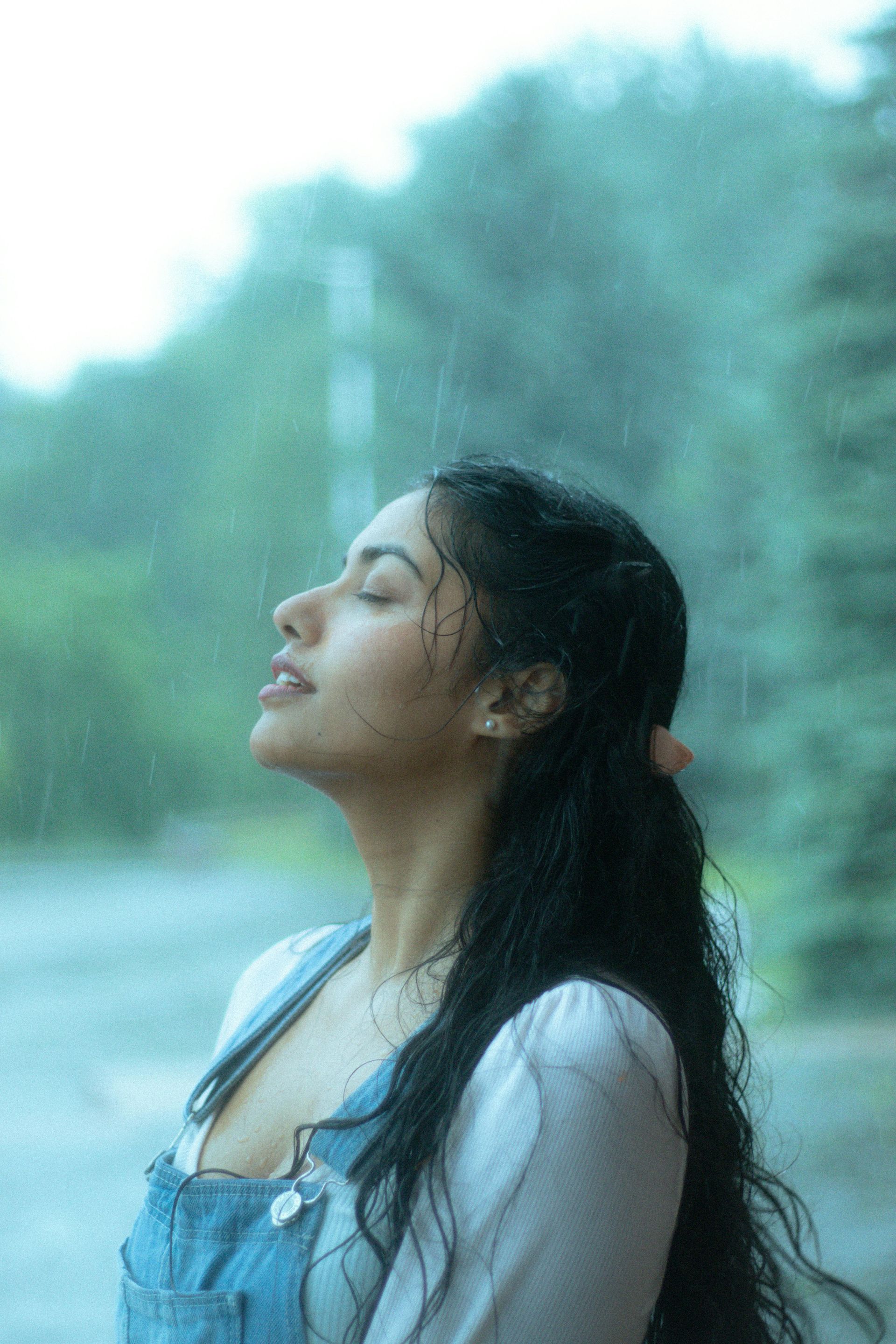 Woman with dark hair in the rain, eyes closed, looking up. Wearing overalls and a white top. Green trees in the background.