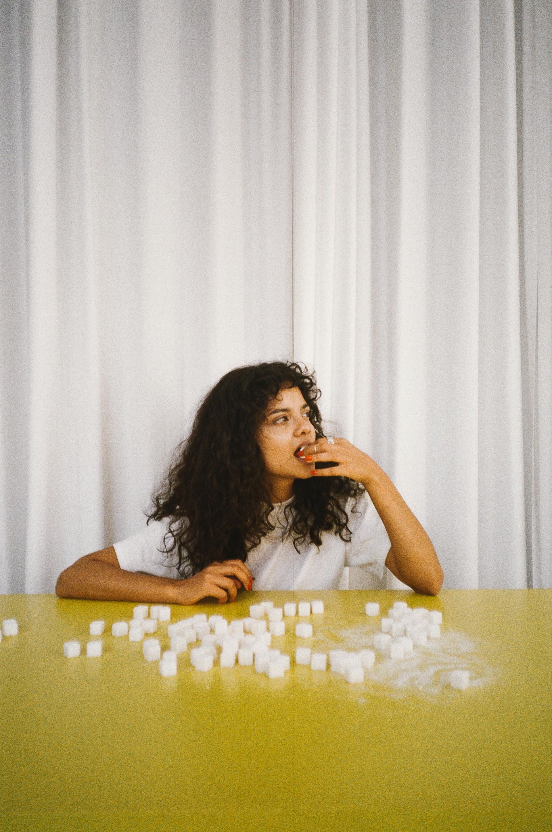 Woman with curly hair eating sugar cubes at a yellow table; white curtain backdrop.