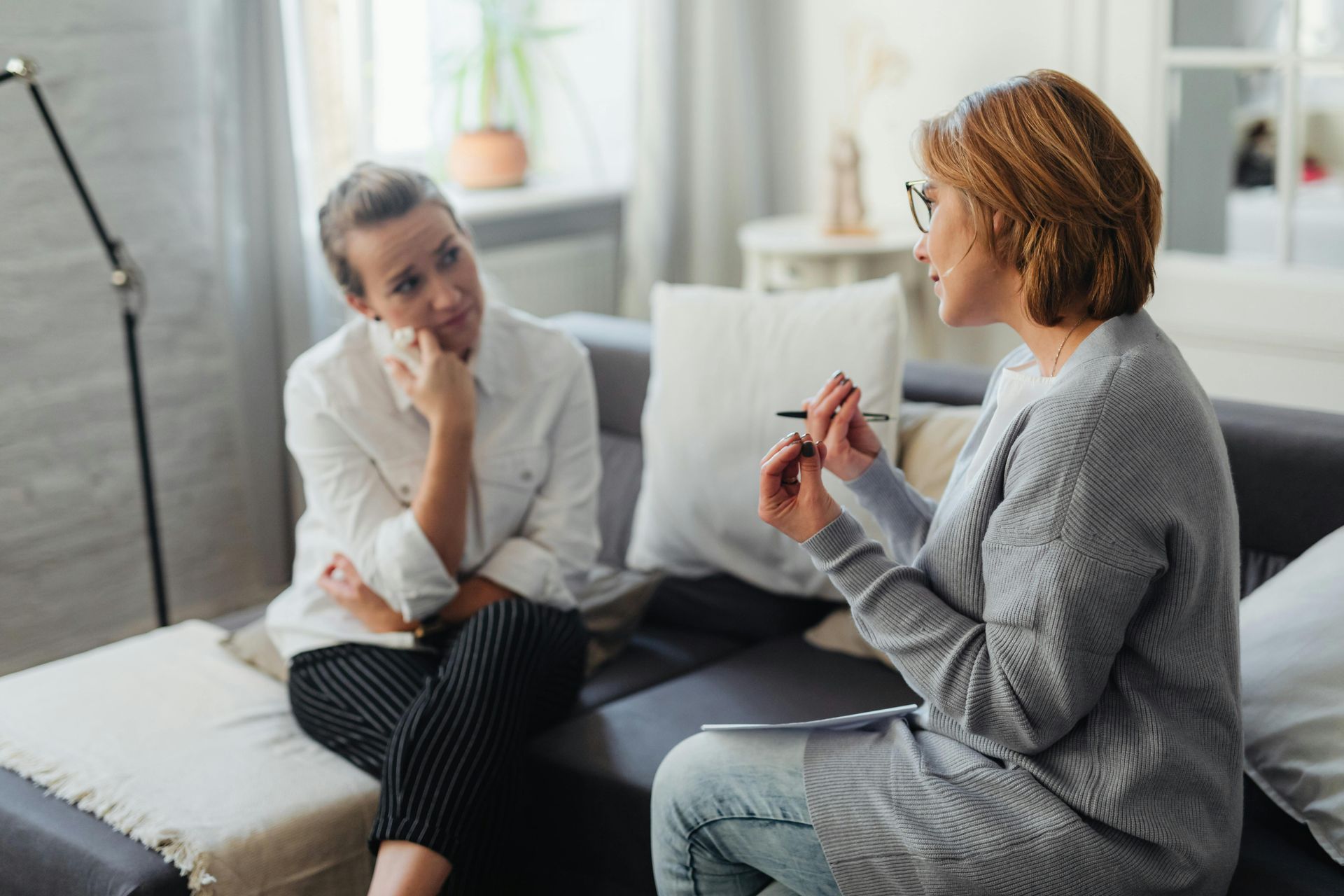 Woman seated on a couch talking to another woman who is holding a pen; living room setting.