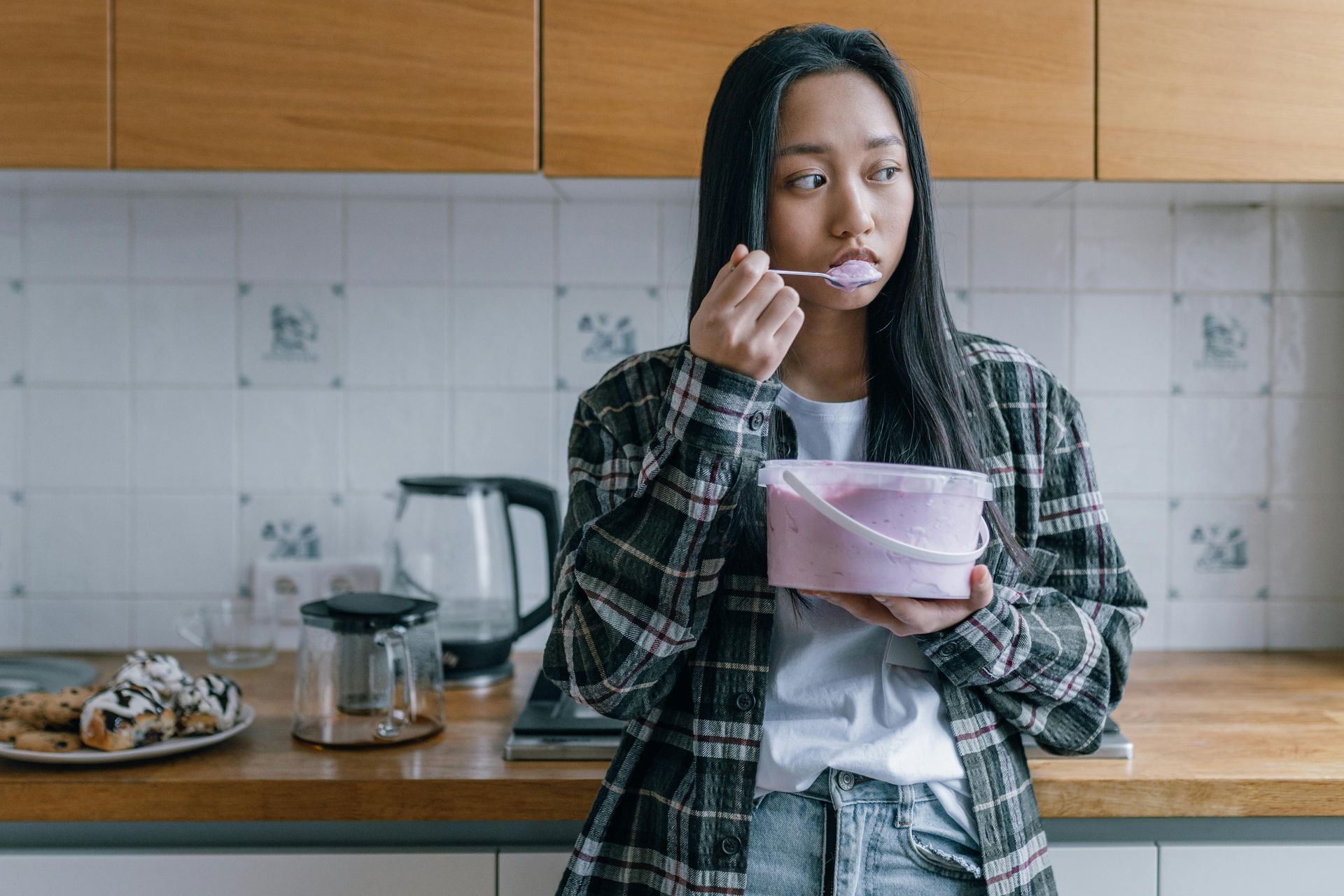 A young woman in a plaid shirt eats ice cream from a tub in a kitchen