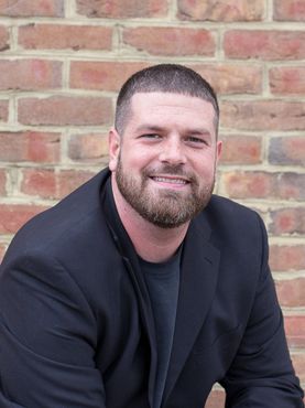 A man with a beard is sitting in front of a brick wall.