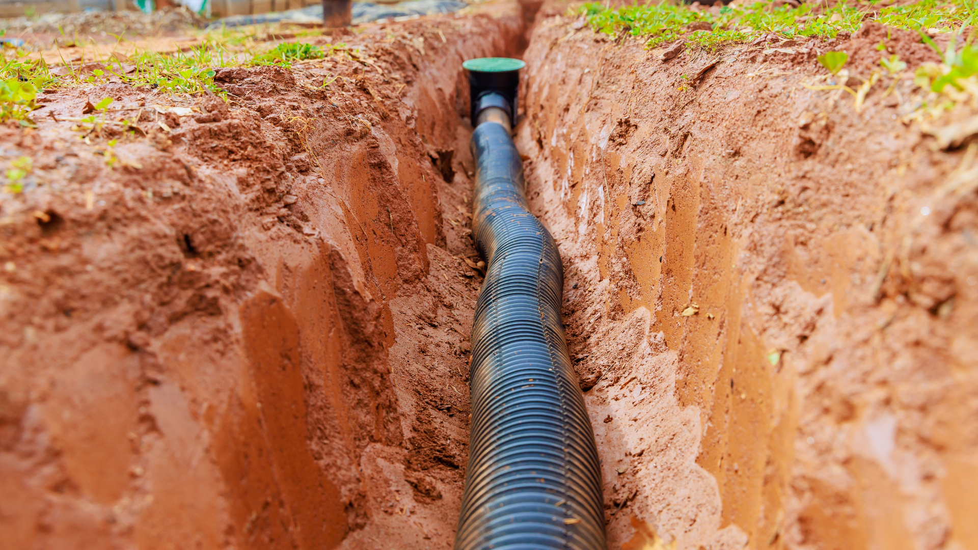 Black corrugated pipe in a trench, surrounded by brown soil and green grass.