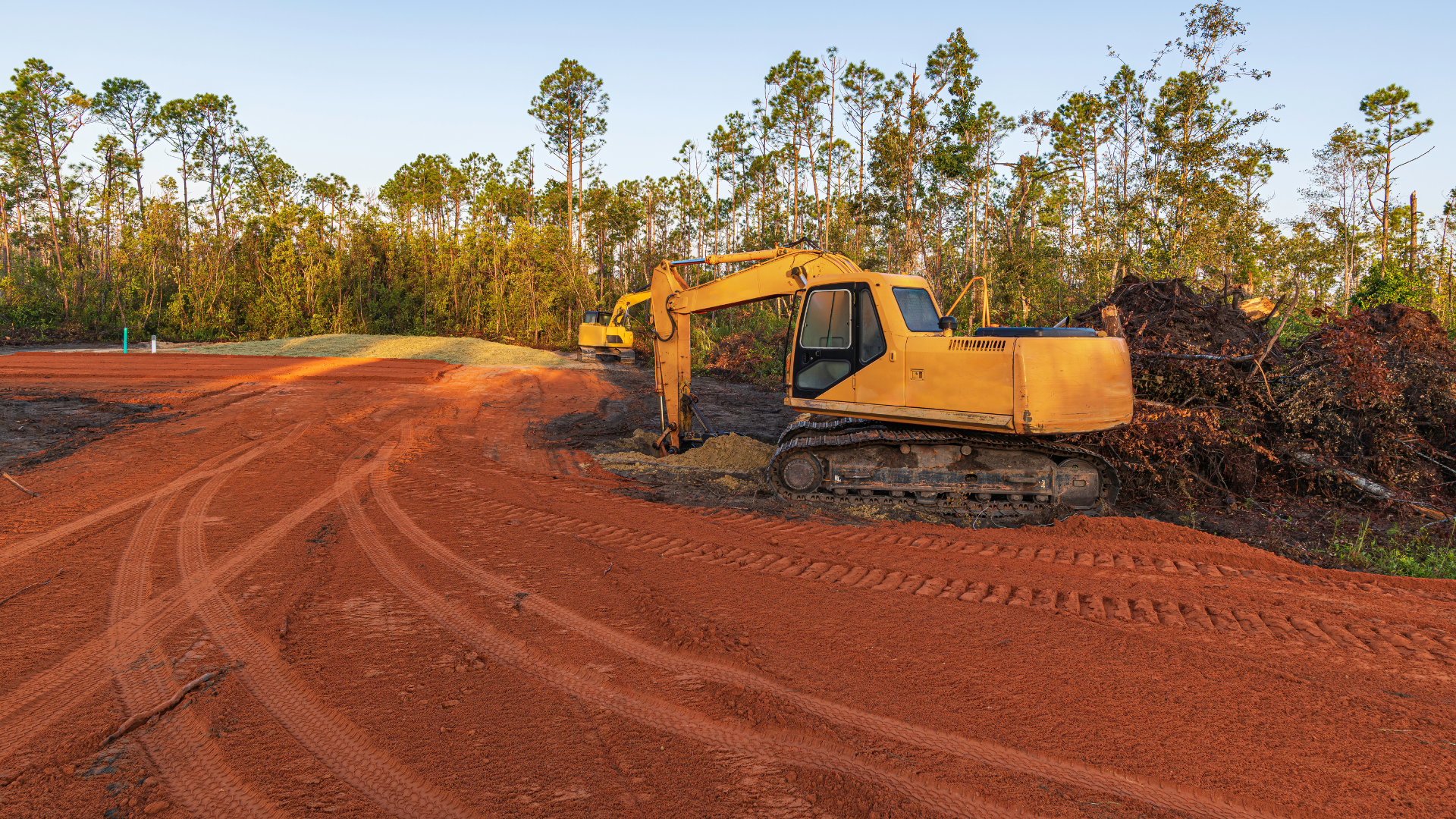 Yellow excavator on red dirt clearing land, trees in background.