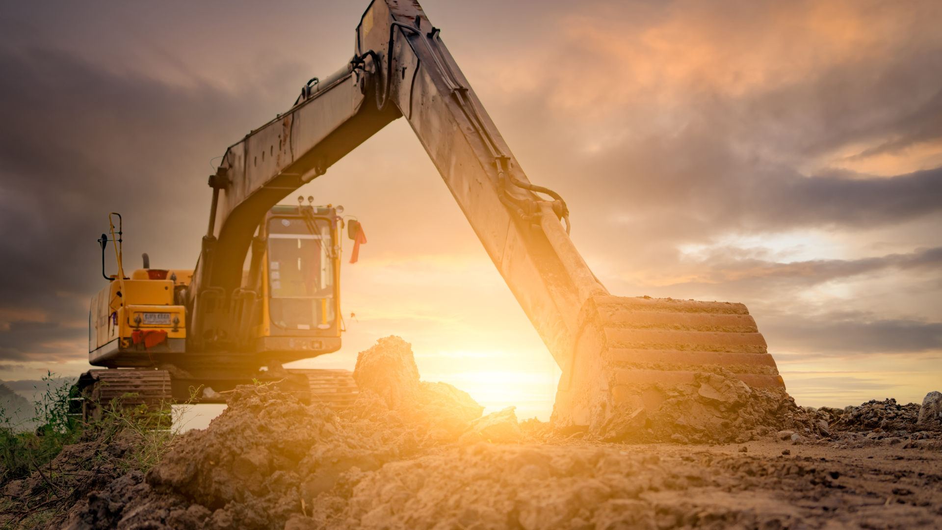 A yellow excavator scoops dirt at a construction site, backlit by a bright sunset.