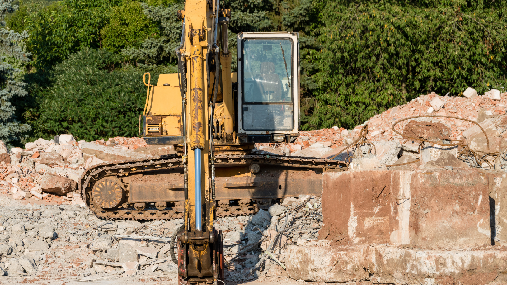 Yellow excavator demolishing a building, surrounded by rubble, with green trees in the background.