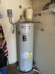 A tall, cylindrical water heater in a corner, with pipes and a pressure tank attached.