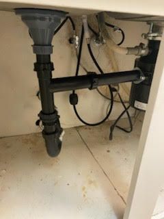 Underside of a sink with plumbing and a garbage disposal. Black pipes, gray drain, white cabinet interior.