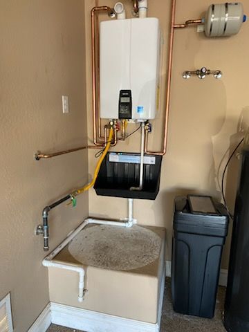 White tankless water heater installed on a beige wall, with copper pipes and a drain pan.