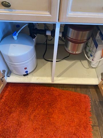 Under-sink cabinet with white water tank and garbage disposal, orange rug visible.