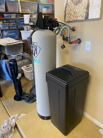 Water softener system in a cluttered utility area. Gray tank and black brine tank next to beige wall.