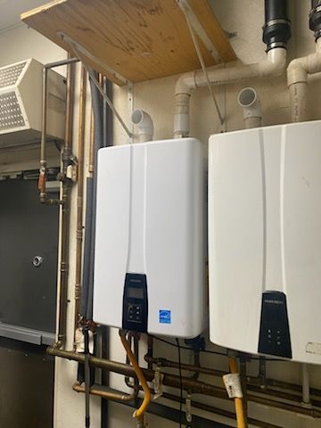 Two white tankless water heaters mounted on a wall with copper pipes and ventilation.