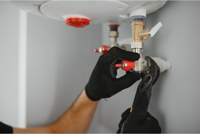 Person with black gloves using a wrench on plumbing near a water heater.