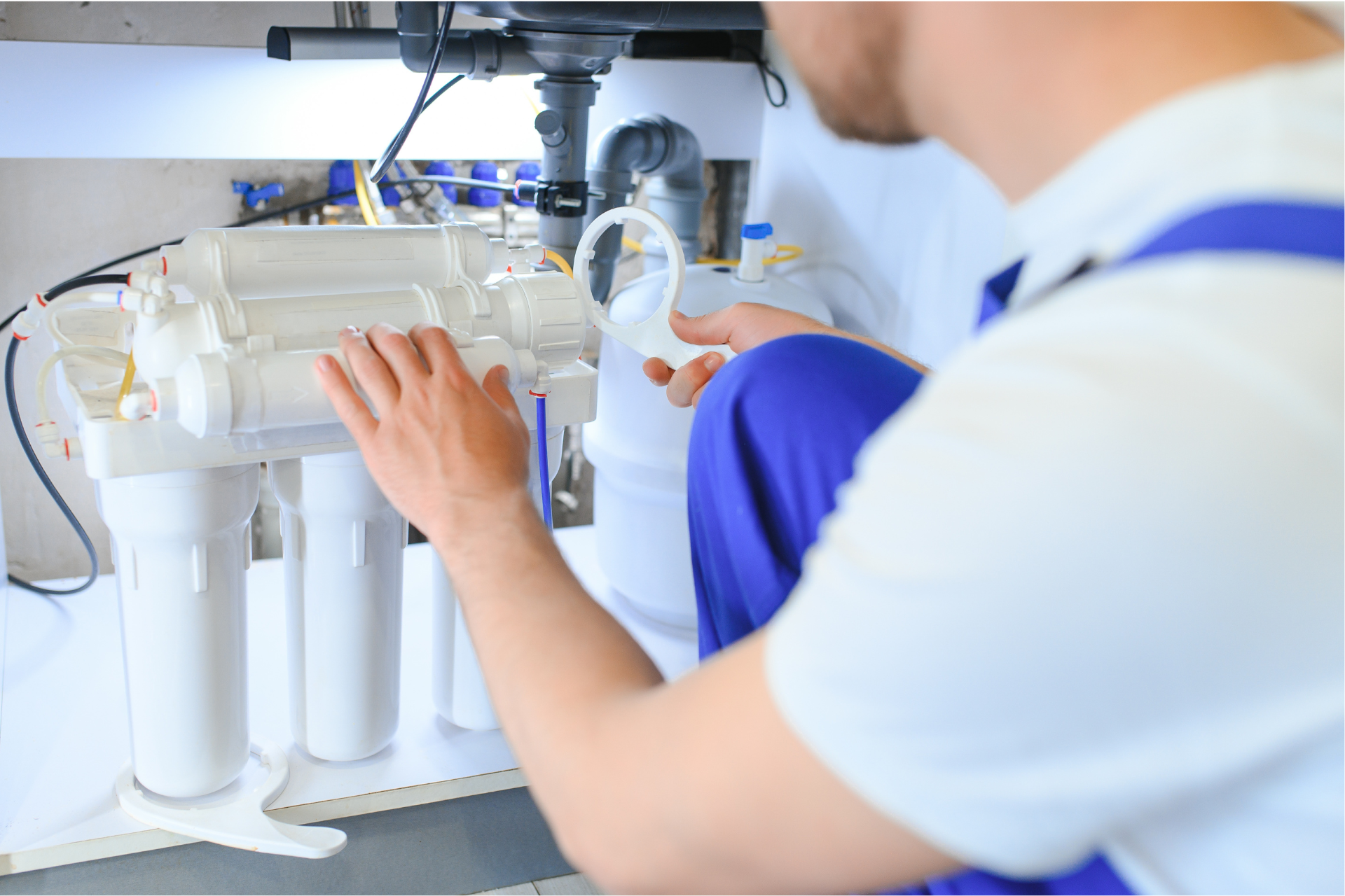 Plumber in blue overalls working on water filtration system under a sink.