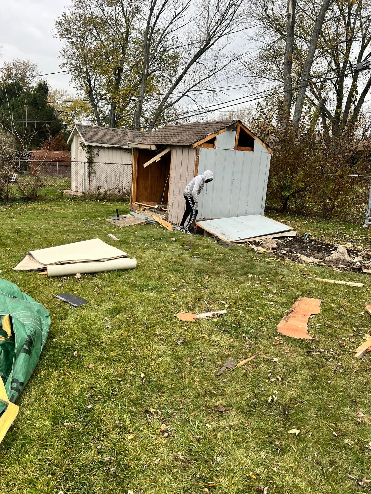 Person dismantling a dilapidated, light blue shed in a grassy backyard. Debris and materials are scattered around.