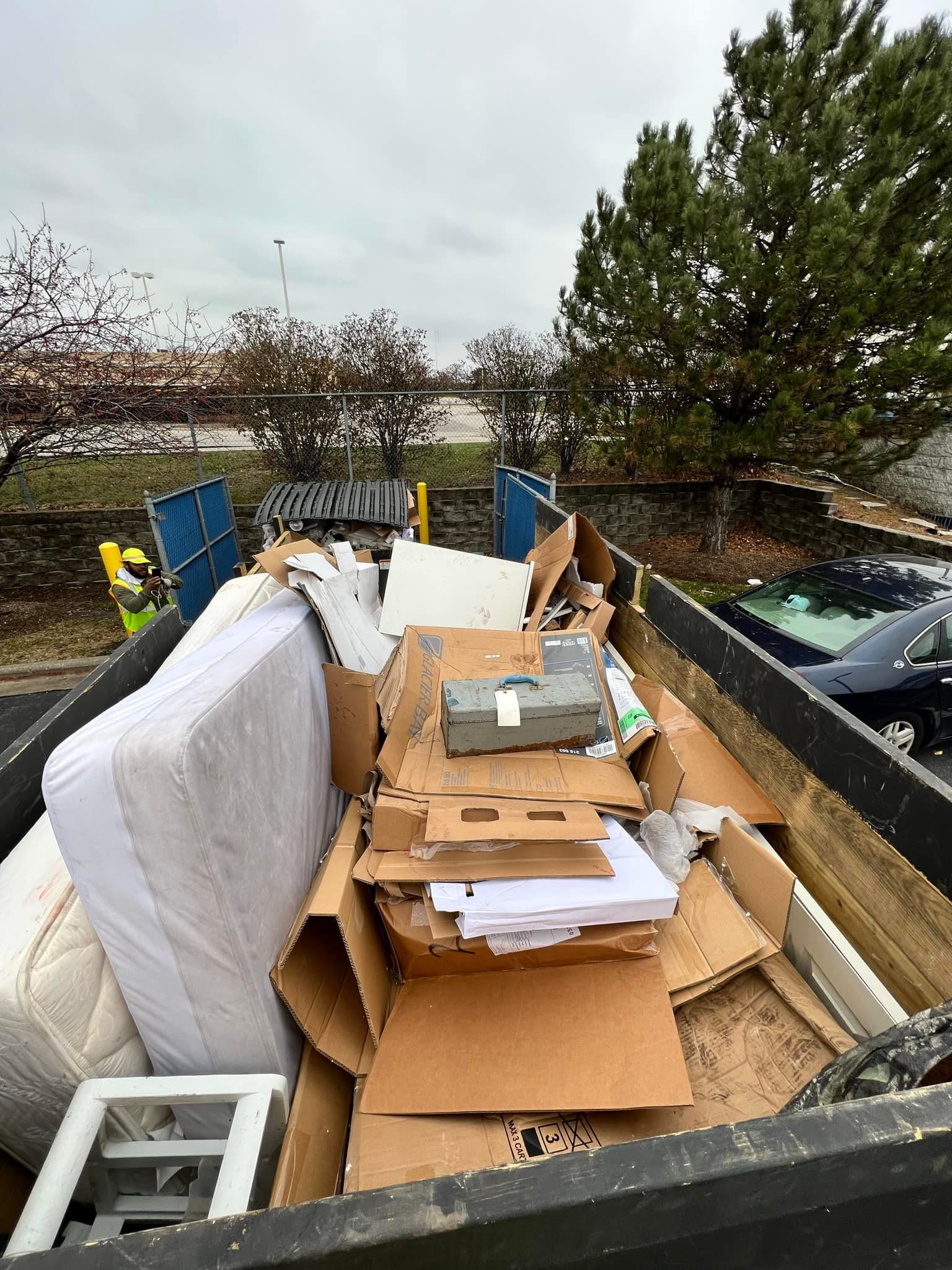 A dumpster filled with trash including cardboard boxes, a mattress, and other debris; set outdoors with trees and a building in the background.