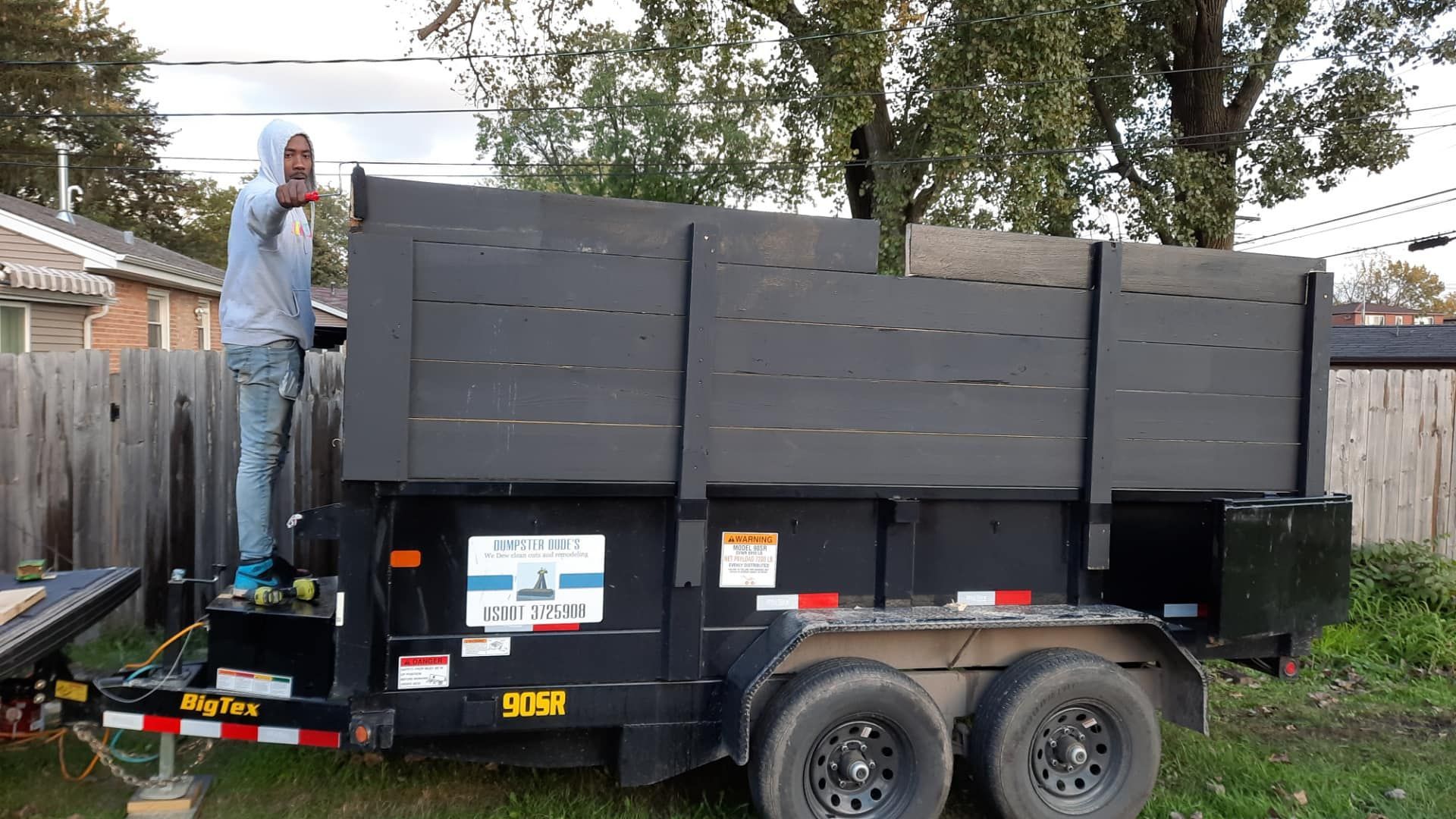 Man in a hoodie standing on a black trailer, gesturing. A wooden fence and houses are in the background.