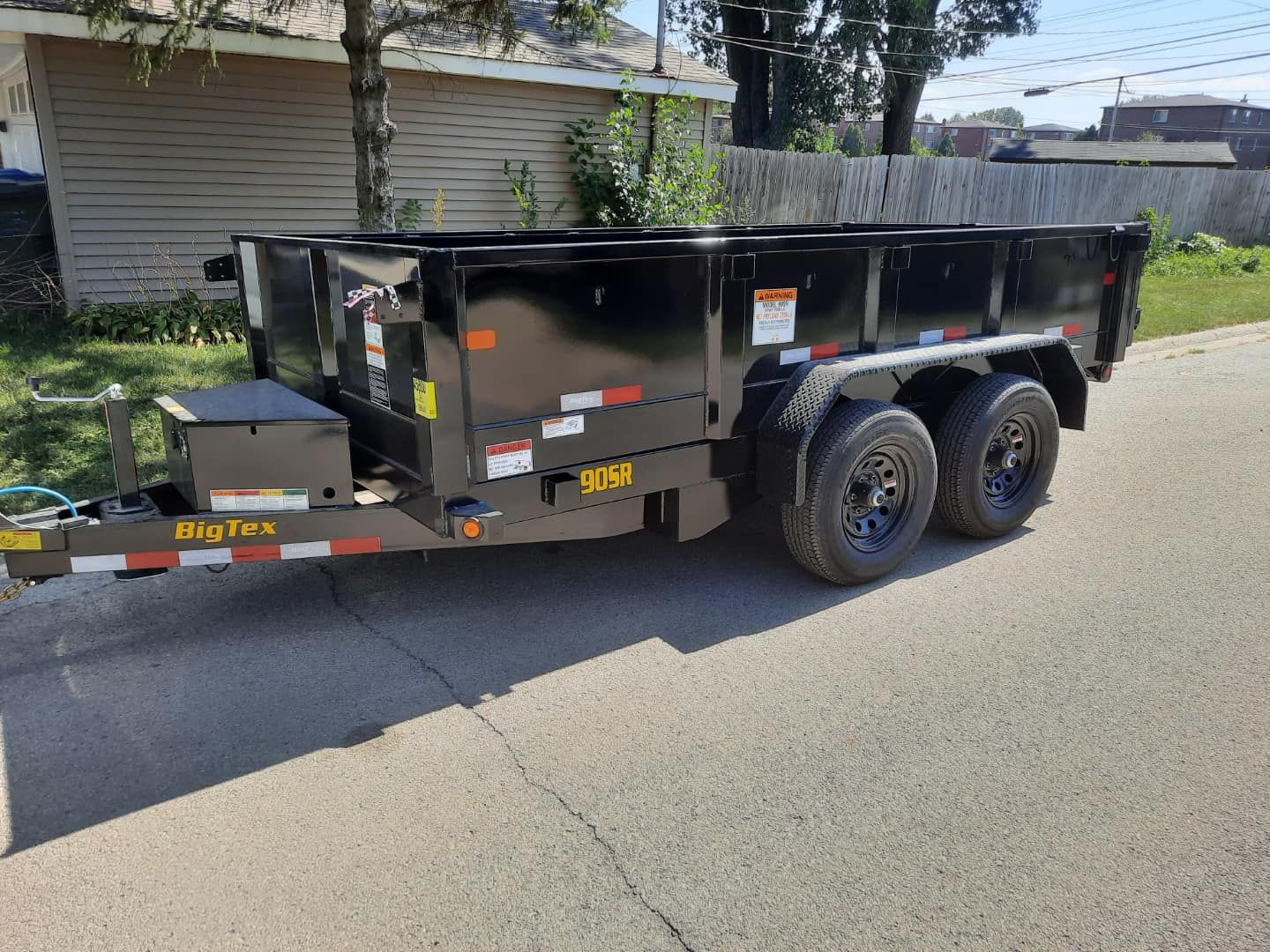 Black Big Tex dump trailer parked on a paved road, with dual wheels and safety reflectors. Background includes houses and trees.