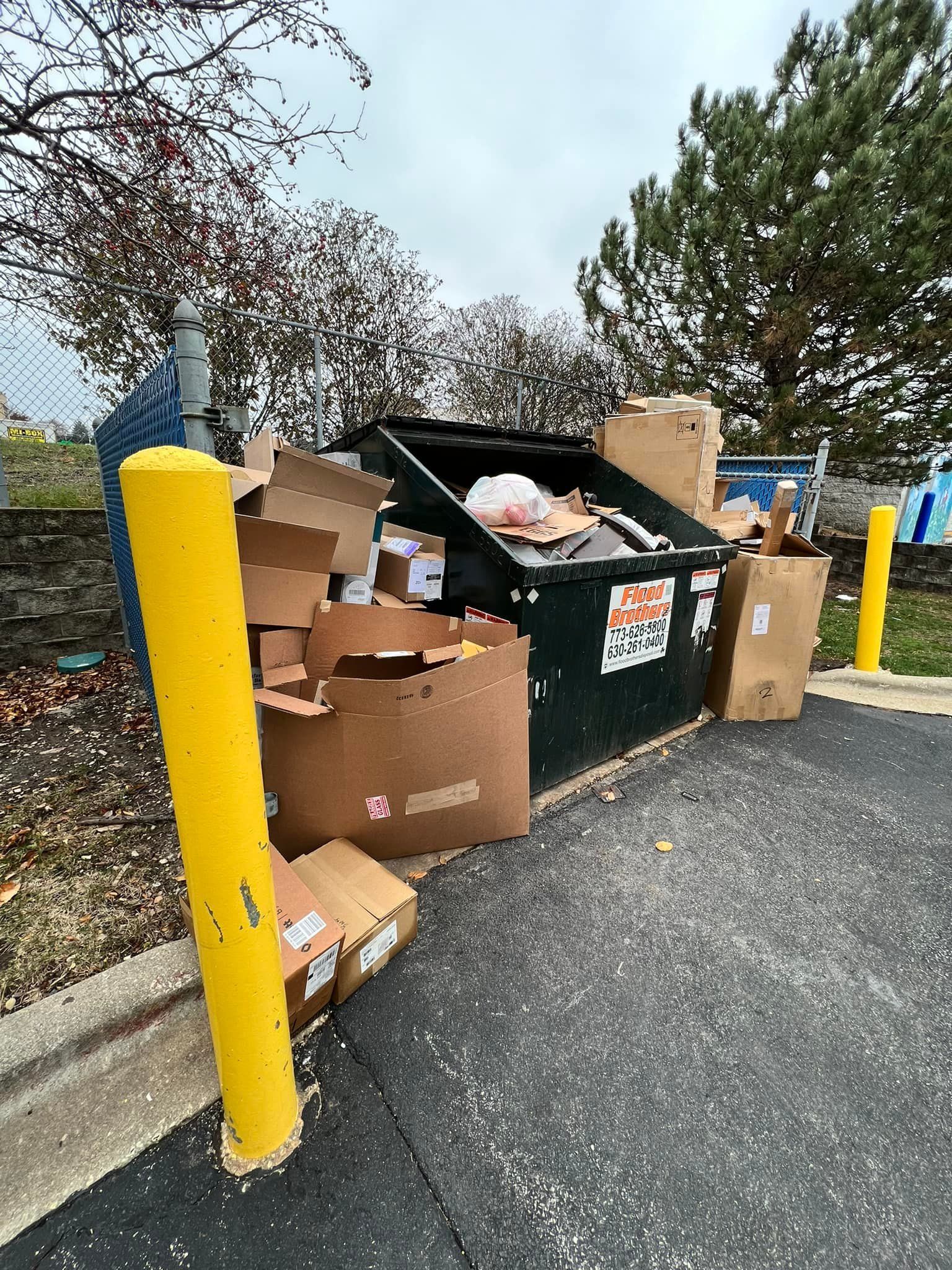 A full dumpster overflowing with trash and cardboard boxes next to a yellow safety post on a paved area.