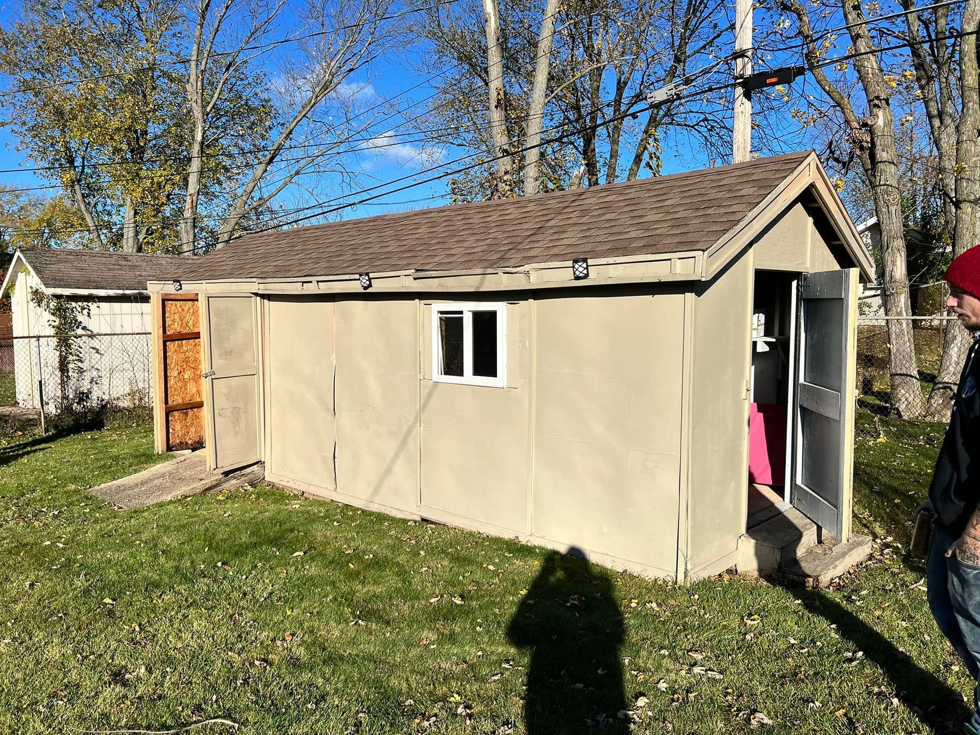 A tan shed with a brown roof, open door, and small window sits on grass. A person is visible to the right.