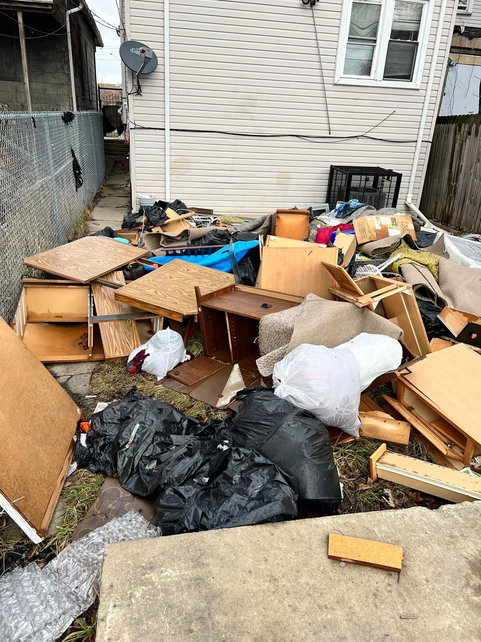 Backyard cluttered with discarded furniture, garbage bags, and debris near a house with a satellite dish.