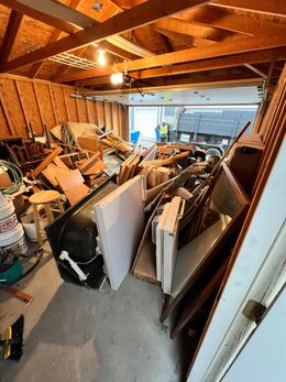 Cluttered garage filled with assorted junk, including doors, furniture, and building materials; garage door open to a street.