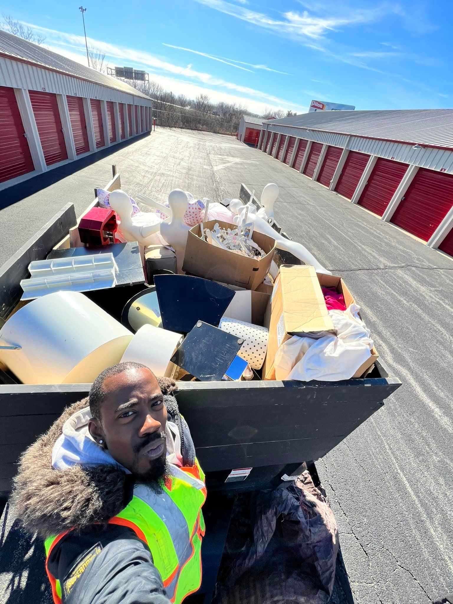 A person in a reflective vest and jacket takes a selfie with a full container of discarded items, outside storage units.