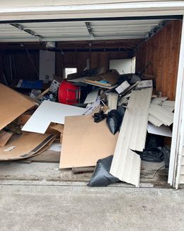 A garage filled with a large pile of debris, including cardboard, wood, and trash bags. A red drum is visible.