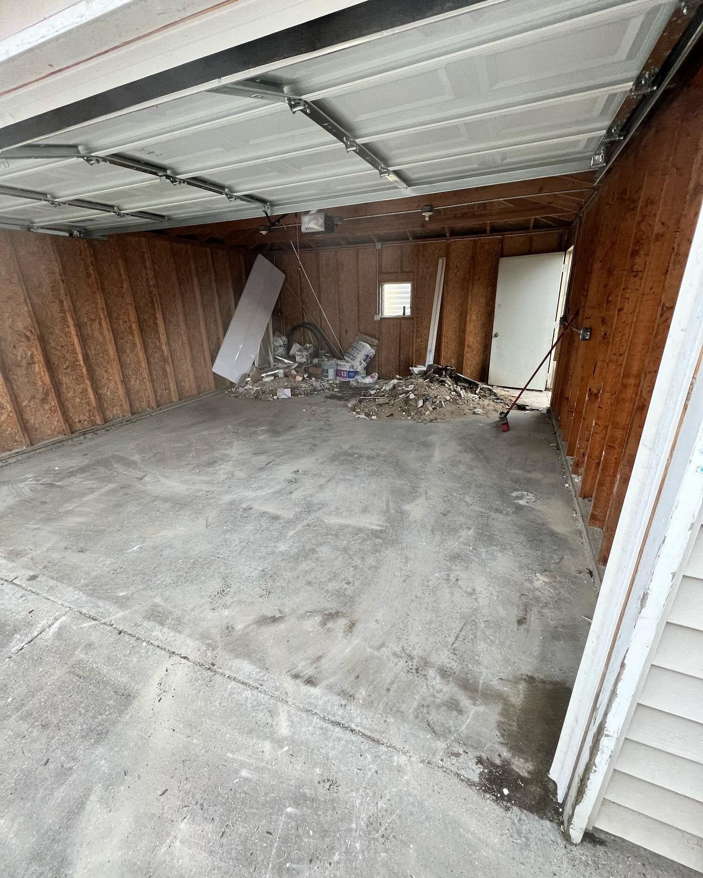 A dirty garage with debris in the center and an open door to the right. Brown wood paneling lines the walls, and the ceiling is white.