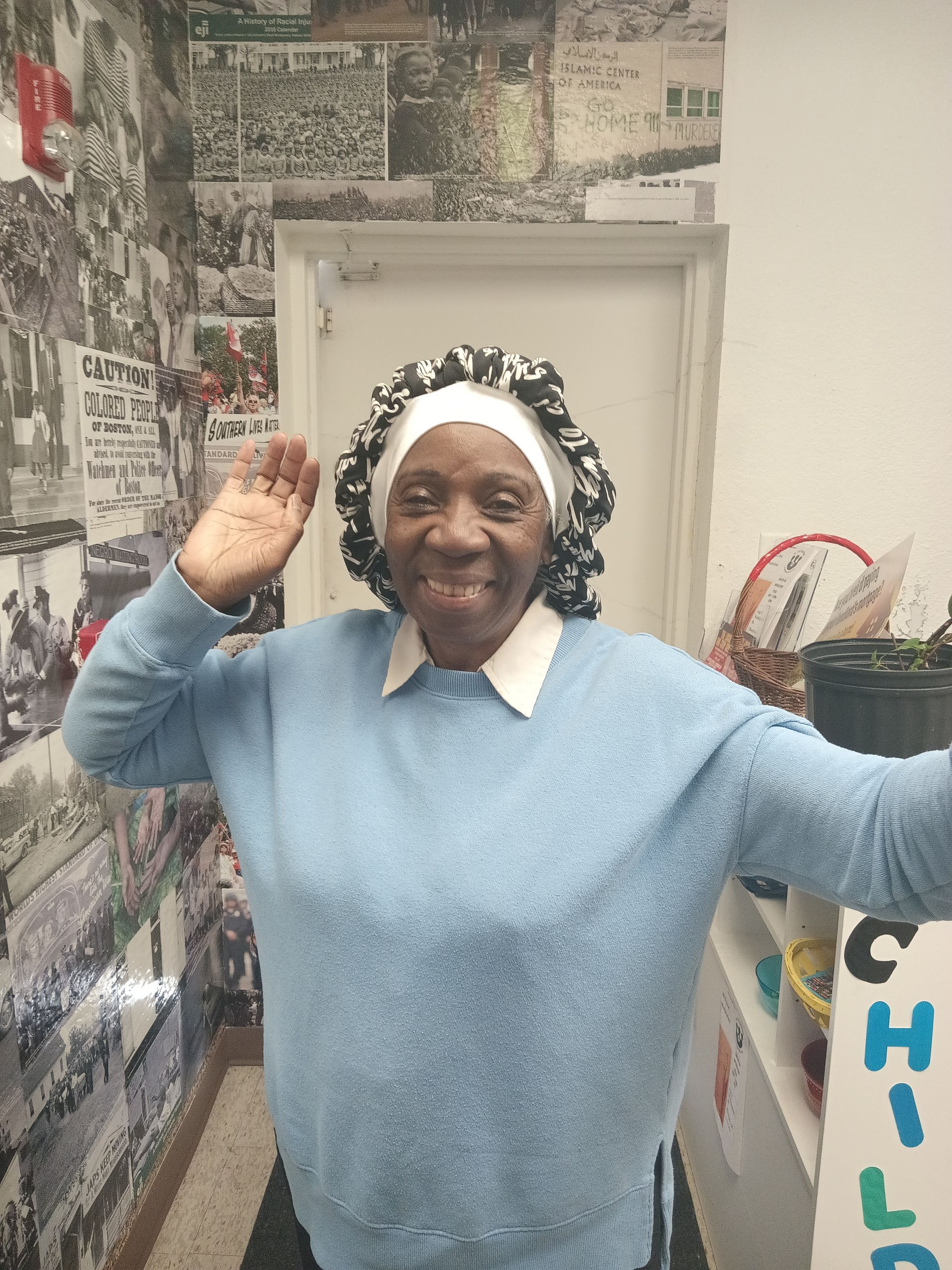 A woman in a blue sweater is standing in a hallway with her arms outstretched.