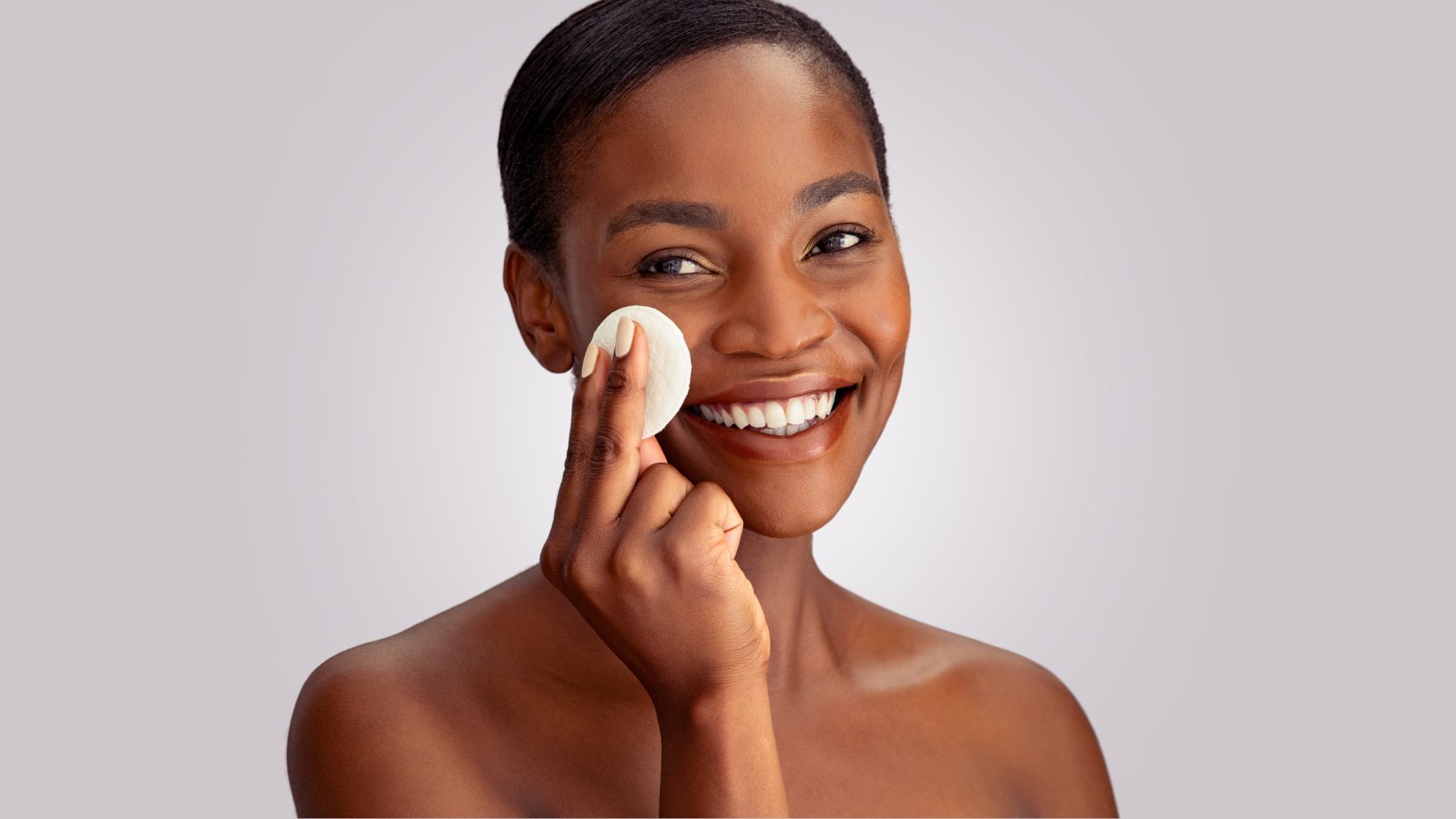 A woman is smiling while cleaning her face with a cotton pad.
