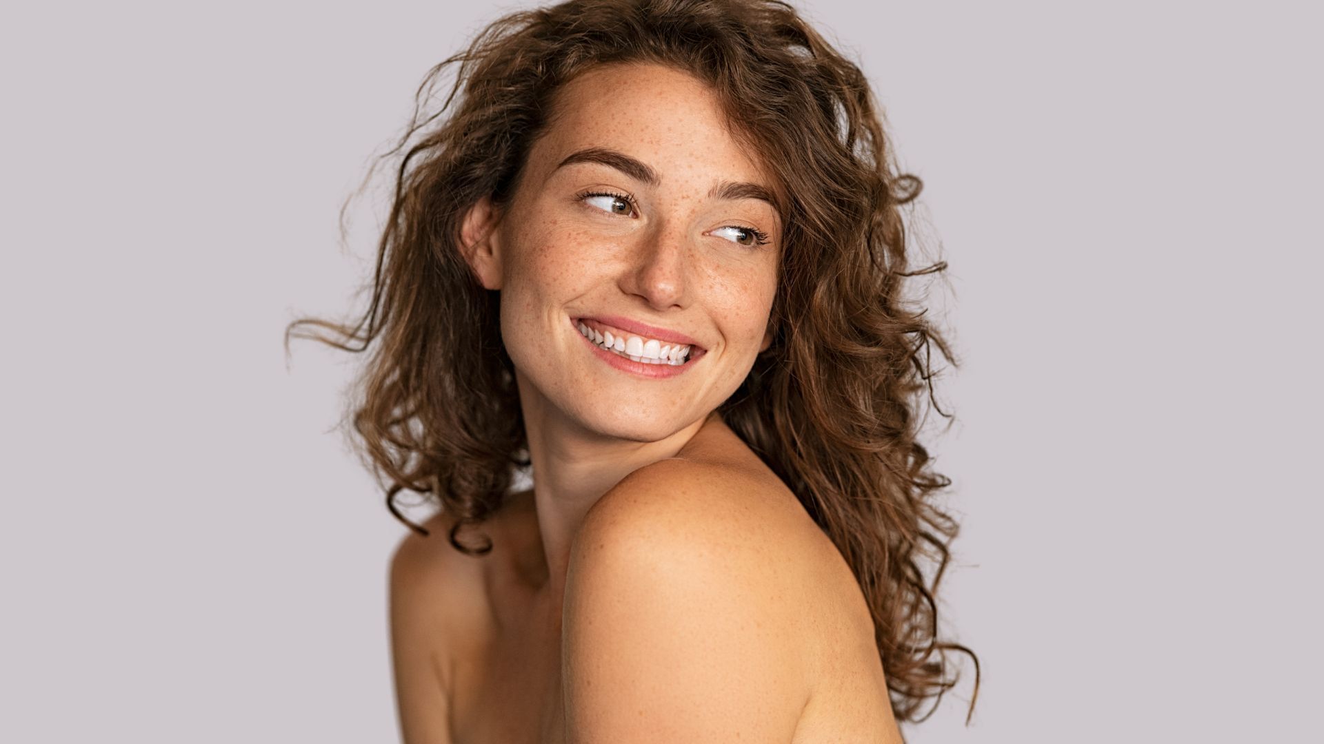 A woman with curly hair is smiling and looking over her shoulder