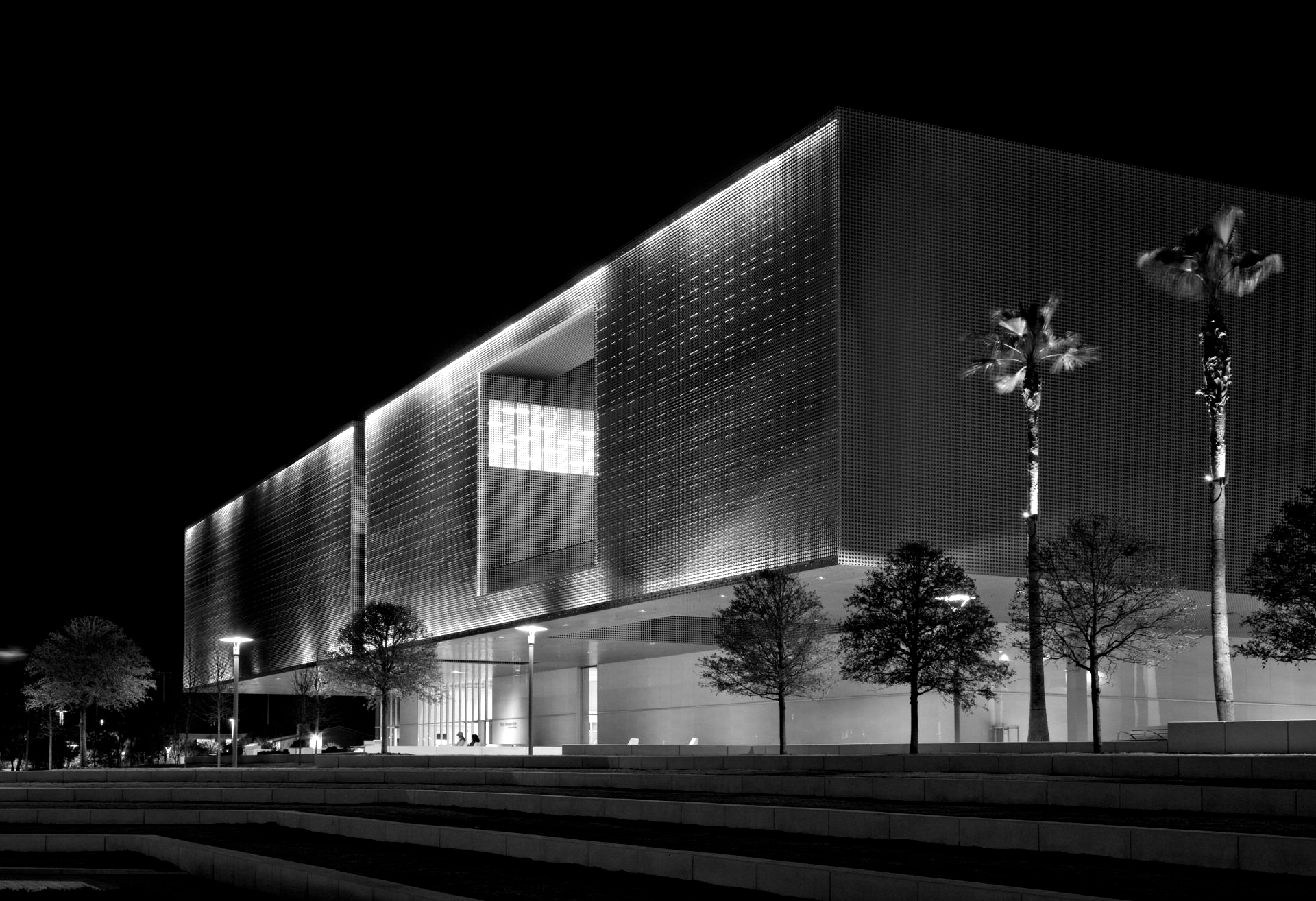 Night view of a modern building with a perforated facade, illuminated by spotlights and featuring palm trees.