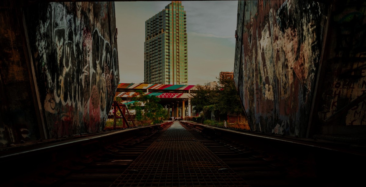 Graffiti-covered tunnel leads to a cityscape with a high-rise building under a cloudy sky.