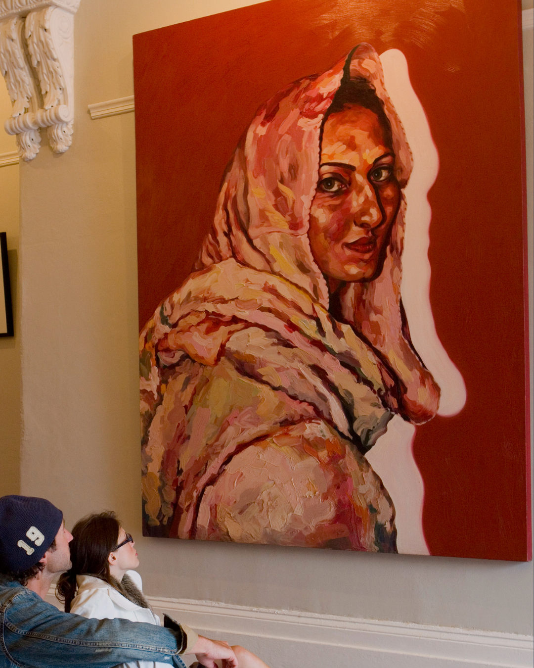 Couple looking at portrait of a woman in a headscarf. Red background, warm tones.