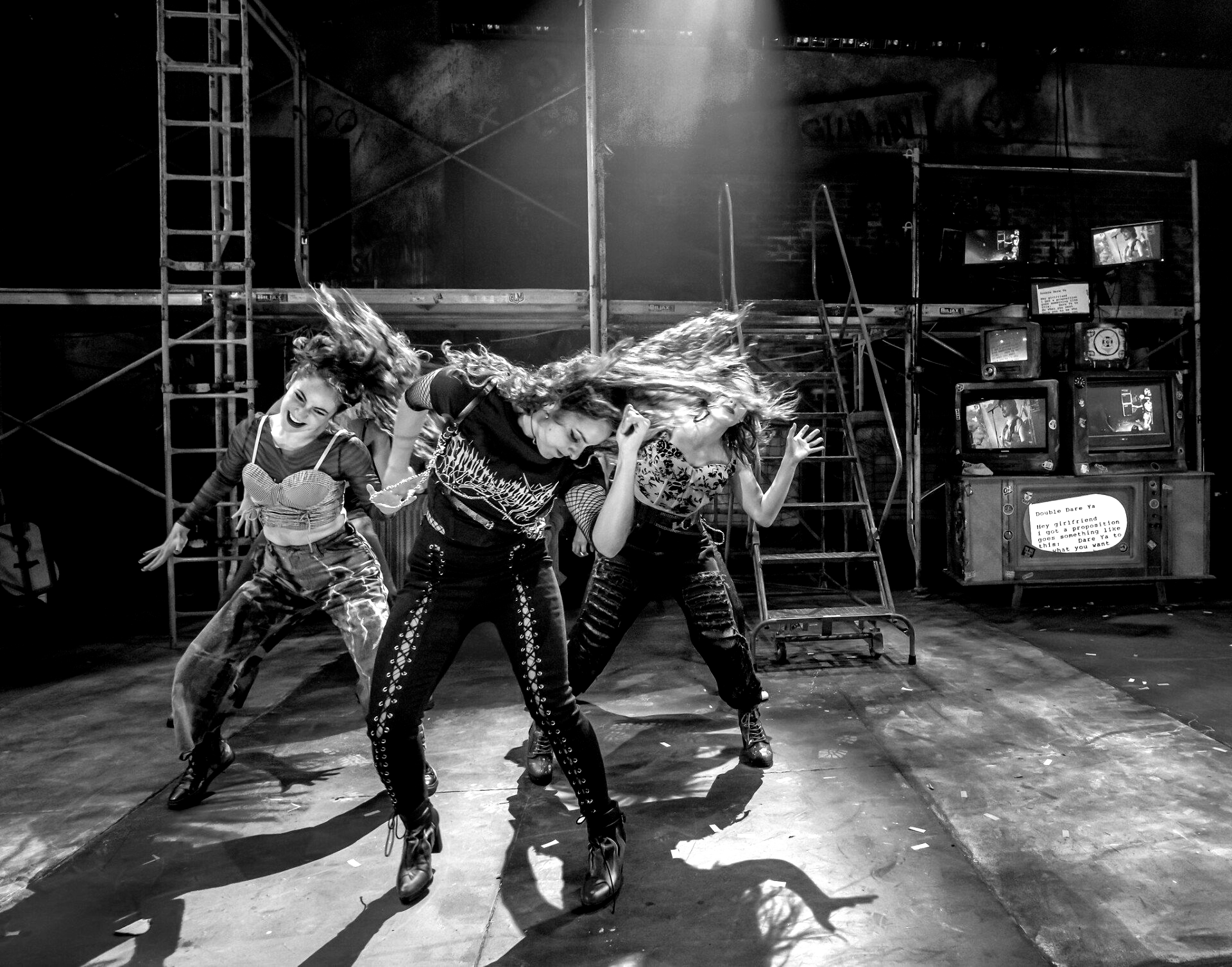 Dancers on a stage, black and white photo. They move energetically, hair flying. Set is industrial, with TVs in the background.